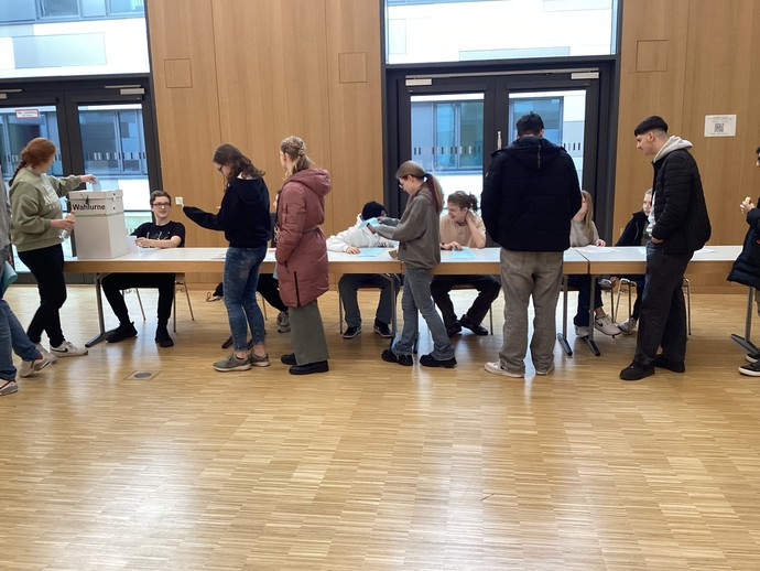 Lernende gehen an einem Tisch mit Wahlurnen und Lernenden vorbei, die dahinter sitzen.