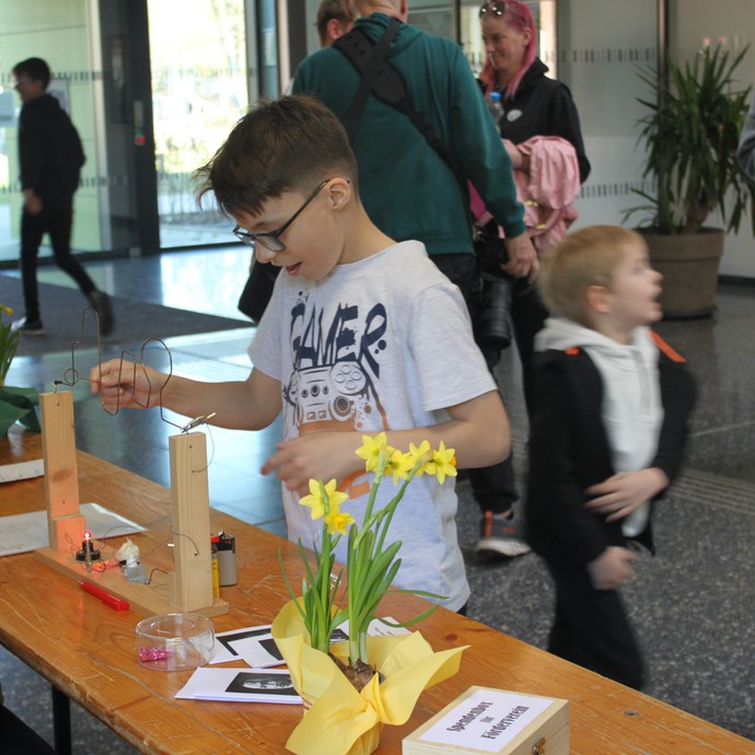 Im Eingangsbereich steht ein Junge an einem selbstgemachten Drahtgeschicklichkeitsspiel und freut sich. Hinter ihm kommen viele Besucher in die Schule (vergrößerte Bildansicht wird geöffnet)