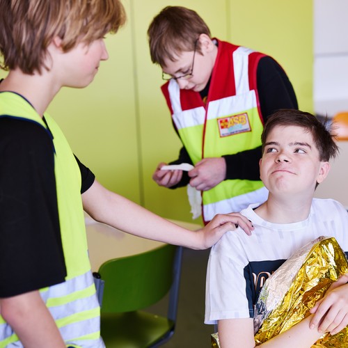 Ein Schüler sitzt in eine Rettungsdecke eingewickelt auf einem Stuhl. Drei Schüler*innen kümmern sich um ihn.