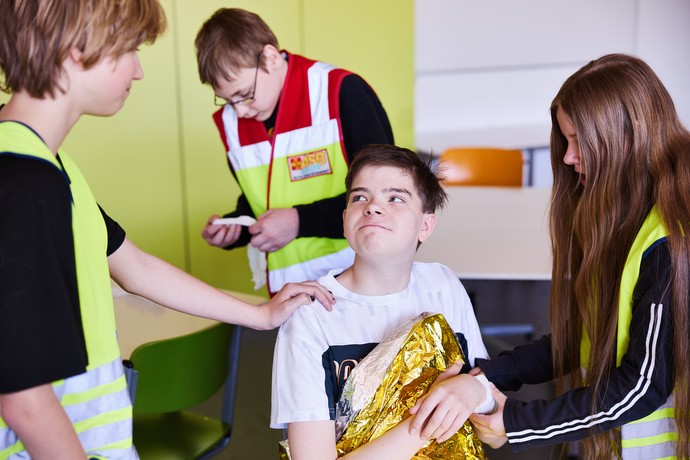 Ein Schüler sitzt in eine Rettungsdecke eingewickelt auf einem Stuhl und drei Schüler kümmern sich um ihn.
