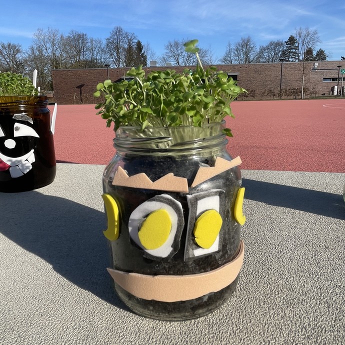 Ein verzierter Glastopf aus dem grüne Pflanzen wachsen (vergrößerte Bildansicht wird geöffnet)