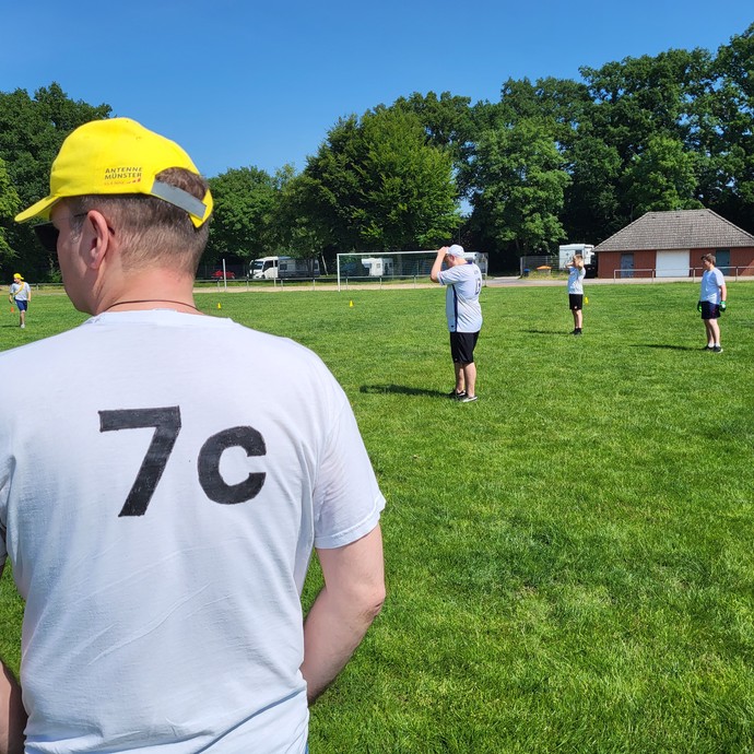 Ein Mann mit gelber Cap und einem Shirt mit Aufdrck 1 c auf dem Rücken und dahinter mehrere Erwachsenen mit Sportkleidung auf dem Feld. (vergrößerte Bildansicht wird geöffnet)