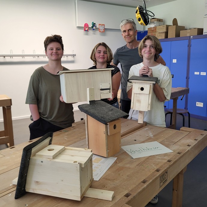 Drei Lernende zeigen mit ihrem Lehrer selbtgebaute Holzvögelhäuser. (vergrößerte Bildansicht wird geöffnet)
