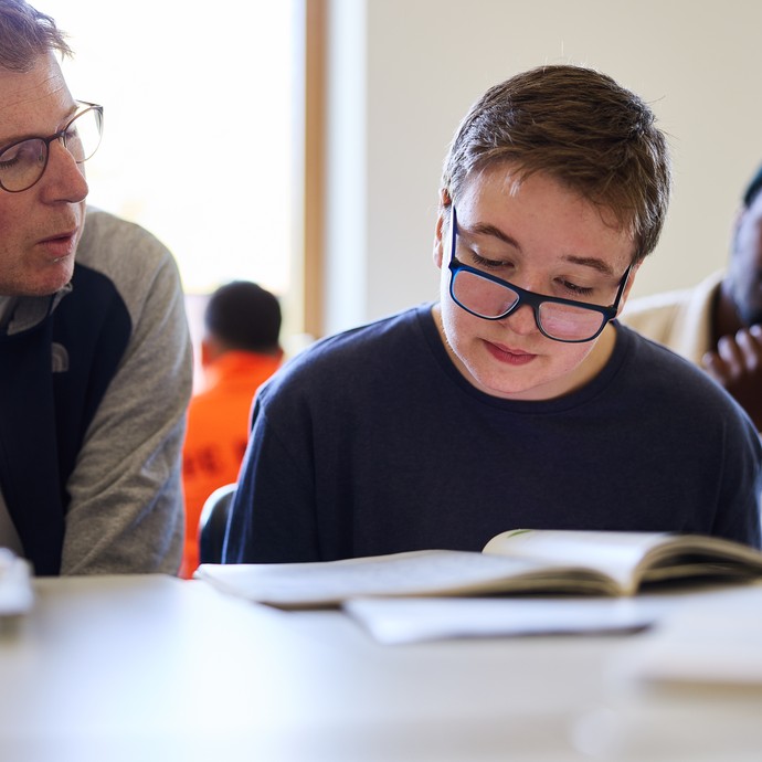 Eine Lerhkraft sitzt neben einem Schüler am Tisch. Im Hintergrund befindet sich ein weiterer Lerndender. (vergrößerte Bildansicht wird geöffnet)