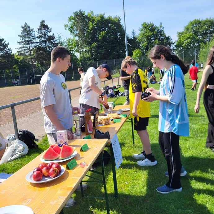 Einige Lernende verkaufen anderen Lernenden im Sporkleidung Waffeln und Obst. (vergrößerte Bildansicht wird geöffnet)