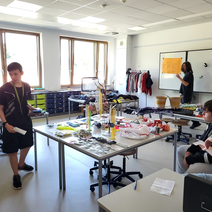 Fünf Lernende befinden sich in einem Klassenzimmer, in dem Ketten, Krawatten und Kleidungsstücke auf den Tischen liegen. (vergrößerte Bildansicht wird geöffnet)