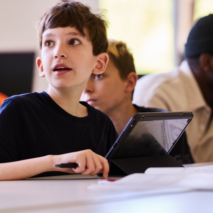 Ein sitzender Schüler blickt aufmerksam nach links. Im Hintergund fefindet sich ein Erwachsener mit einem weiteren Lernenden. (vergrößerte Bildansicht wird geöffnet)