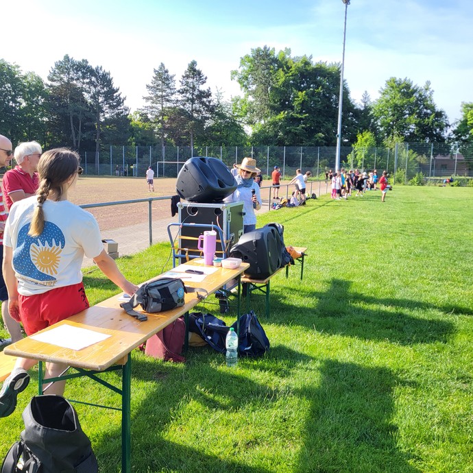 Drei Personen stehen zwischenFußbalfeld, Holzklappbank und Lautsprecherbox. Im Hintergrund sind weitere Personen zu sehen. (vergrößerte Bildansicht wird geöffnet)