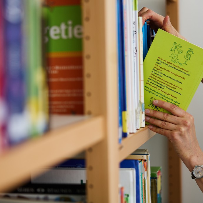 Ein Bücherregal aus dem eine Hand sich ein Buch nimmt. (vergrößerte Bildansicht wird geöffnet)
