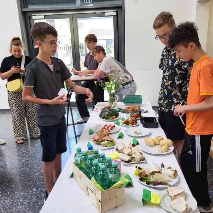 Drei Schüler stehen an einem Buffet. Dahinter sieht man weitere Personen, die etwas essen und sich unterhalten. (vergrößerte Bildansicht wird geöffnet)