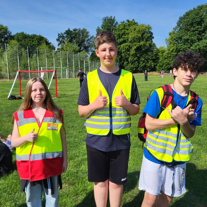 Drei Lernende stehen mit Warnwesten und Daumen hoch vor dem Fußballfeld mit weiteren Personen. (vergrößerte Bildansicht wird geöffnet)