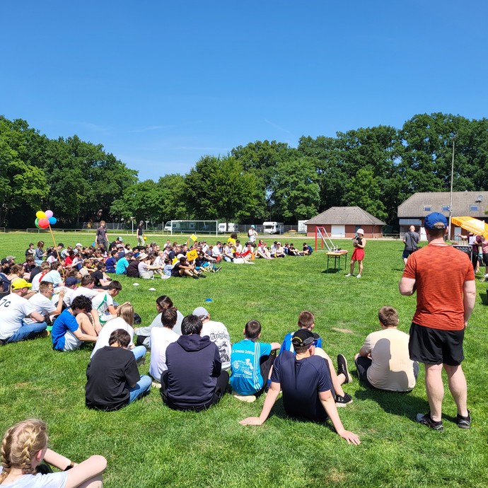 Viele Personen sitzen auf auf dem Rasen des Sportplatzes und schauen zu einer Frau, die in der Mitte steht. (vergrößerte Bildansicht wird geöffnet)
