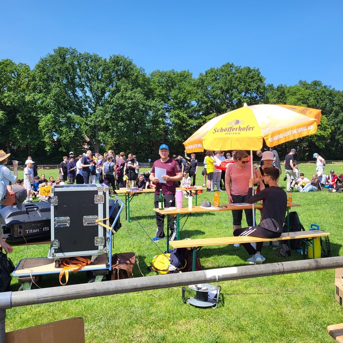 Auf dem Sportplatz stehen Holzbänke und Tische. Auf dem Feld sind viele Personen zu sehen. (vergrößerte Bildansicht wird geöffnet)