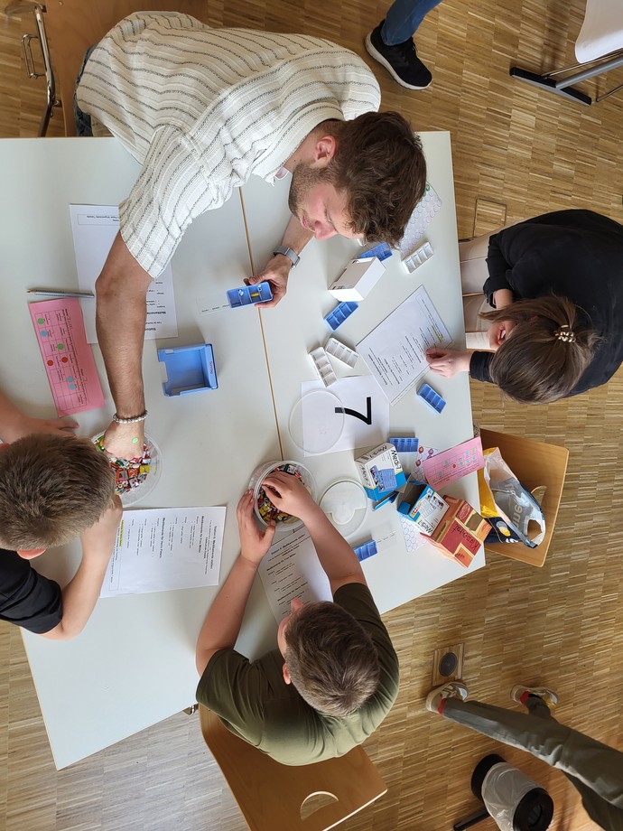 Vier Lernende sitzen an einem Tisch und sortieren dort stellvertretend Süßigkeiten in Meikamentendosierer.
