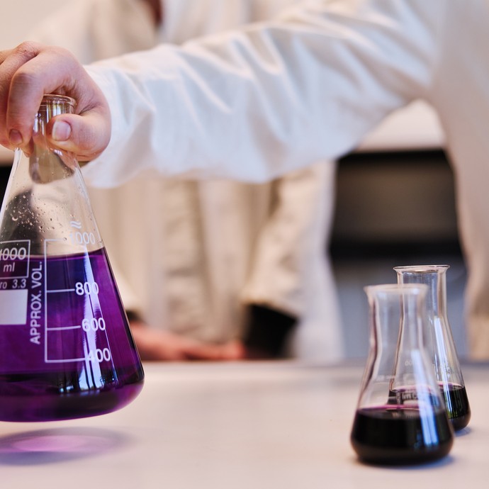 Eine Hand stellt eine große Flasche mit lila Flüssigkeithin. In zwei weiteren kleinen Gefäßen, die daneben stehen, ist die selbe Flüssigkeit. (vergrößerte Bildansicht wird geöffnet)