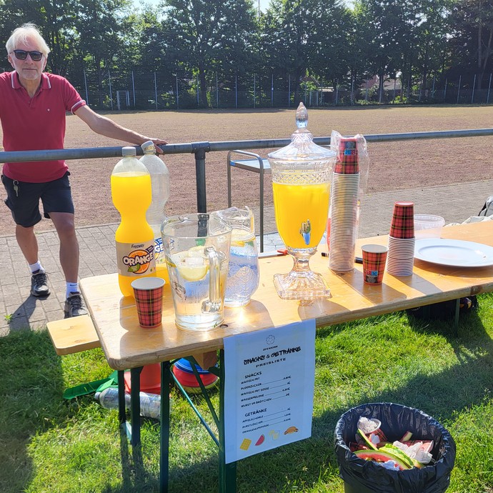 Getränke auf einem Holzklapptisch und dahinter steht ein Erwachsener in Sportkleidung. (vergrößerte Bildansicht wird geöffnet)