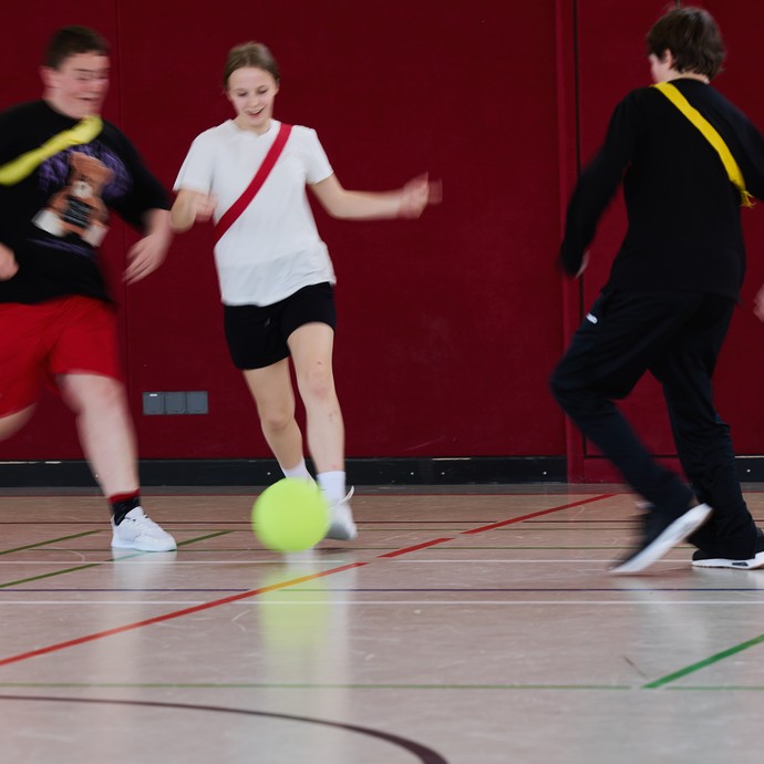 Drei Lernende spieln in der Halle Fußball. Im Hintergrund sieht man die Schiedsrichterin. (vergrößerte Bildansicht wird geöffnet)