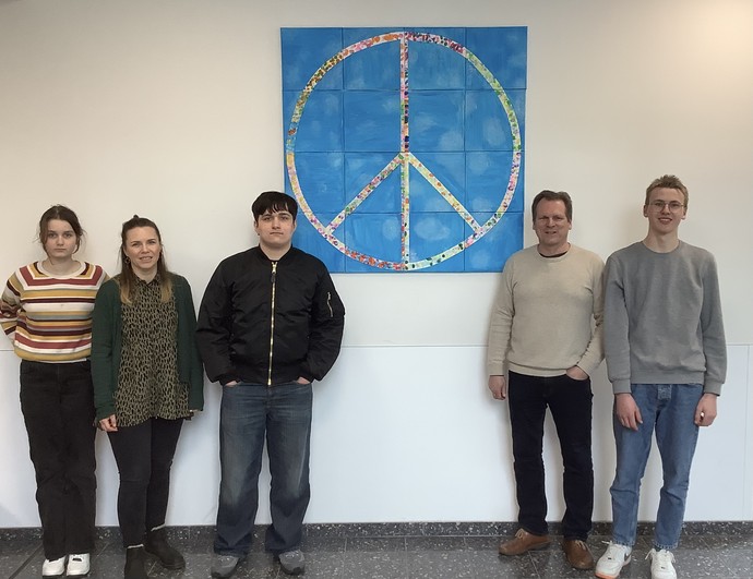Zwei Schülerinnen, zwei Schüler und ein Lehrer stehen vor dem selbstgestalteten Peacezeichen, das an einer Wand hängt.