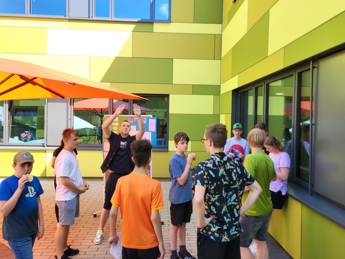 Elf Lernende stehen neben dem Schulgebäude unter einem Sonnenschirm. Einige von ihnen essen etwas.