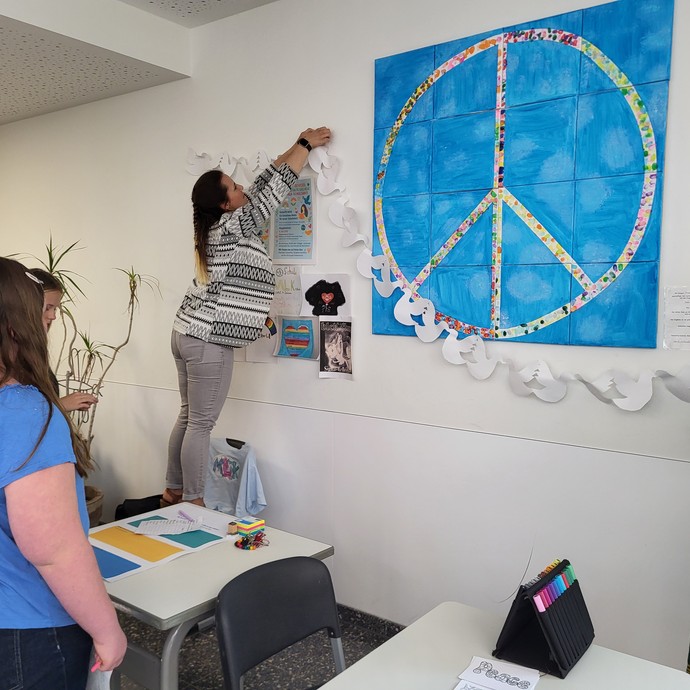 Eine Lehrerin hängt unter ein Bild mit Peacezeichen aus Papier augeschnittenen Friedenstauben. Drei Schülerinnen stehen daneben. (vergrößerte Bildansicht wird geöffnet)