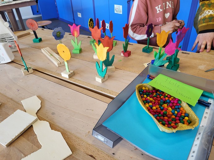 Auf einem Tisch stehen aus Holz gearbeitete und angemalte Frühlingsblumen neben einem Kob mit Perlen.