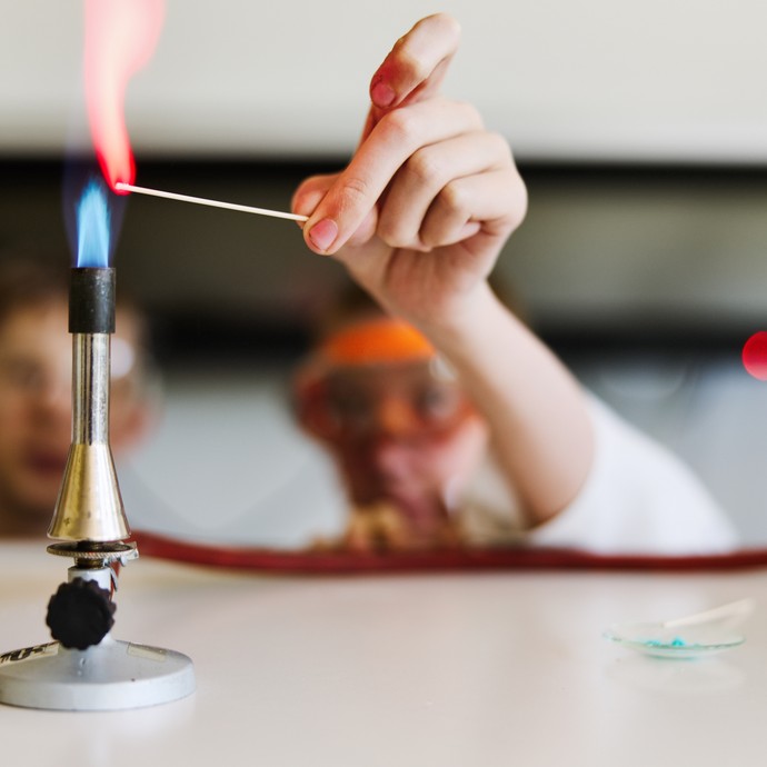 An einen Bunsenbrenner wird ein Holz entzündet. Im Hintergrund sieht man verschwommen zwei Personen mit Schutzbrille. (vergrößerte Bildansicht wird geöffnet)