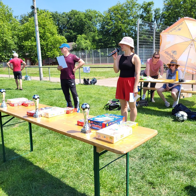 Eine Frau mit Mikrofon steht mit einem Mann hinter dem Tisch auf dem die Pokale stehen und verkündet die Sieger. (vergrößerte Bildansicht wird geöffnet)