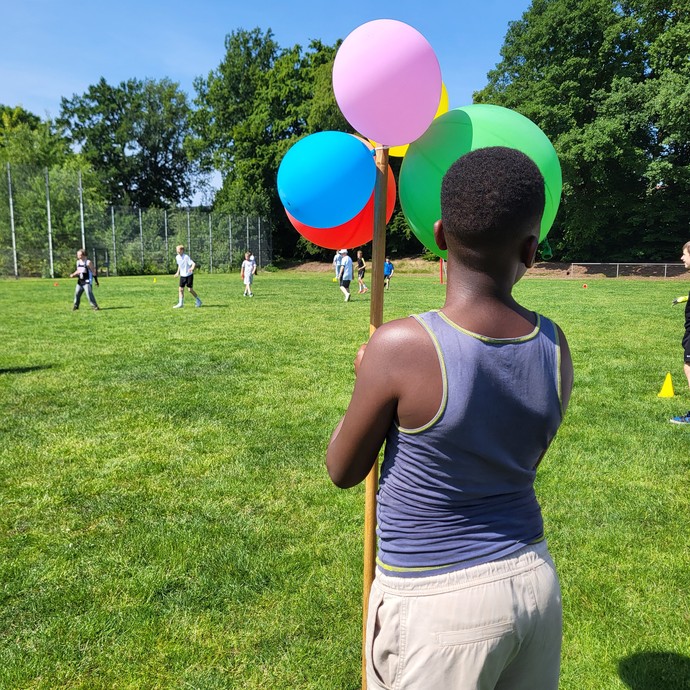 Ein Junge hält eine Holzstange, an der 5 Luftballons gebunden sind. Im Hintergrund sieht man Fußballspieler auf Rasen. (vergrößerte Bildansicht wird geöffnet)