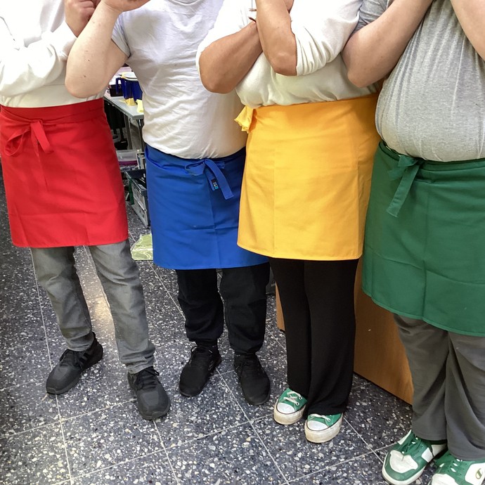 Ein paar Lernende mit bunten umgebundenen Schürzen, von denen man das Gesicht aufgrund des Bildausschnittes, nicht sieht. (vergrößerte Bildansicht wird geöffnet)