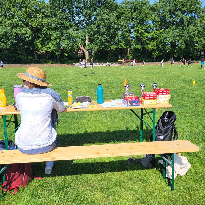 Eine Frau mit Strohhut sitzt auf einer Holzbank. Auf dem Tisch dahinter stehen Pokale. Im Hintergrund befindet sich das Fußballfeld mit Spiielenden. (vergrößerte Bildansicht wird geöffnet)