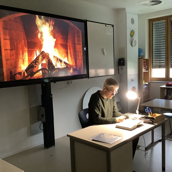 Ein Lehrer liest am Pult. Hinter ihm ist ein Bildschrimpräsentation mit Kaminfeuer zu sehen. (vergrößerte Bildansicht wird geöffnet)