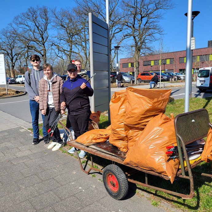 Vier Lernende stehen mit Abfallgreifern hinter einem Wagen zum Anschieben auf dem 3 1/2 volle Müllsäcke liegen. (vergrößerte Bildansicht wird geöffnet)