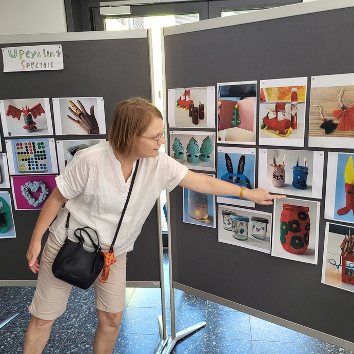 Eine Lehrerin zeigt auf ein Foto, welches mit weiteren Fotos von Werken und der Aufschrift Upcycling auf einer Stellfläche hängt. (vergrößerte Bildansicht wird geöffnet)