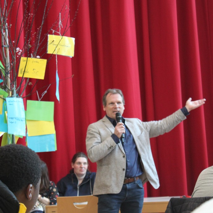 Ein Lehrer steht vor einem roten Vorhang und hebt die Hand nach oben. (vergrößerte Bildansicht wird geöffnet)